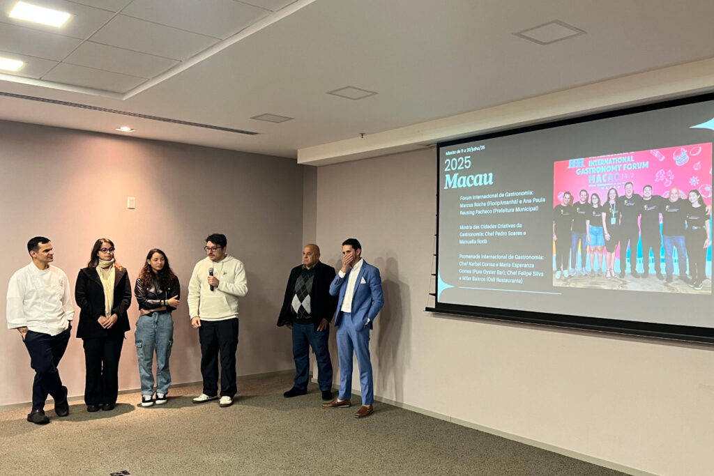 Seis pessoas em pé diante de tela projetada que exibe informações sobre o Fórum Internacional de Gastronomia 2025 em Macau.